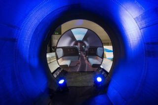 Diseño de interiorismo de la cápsula de HTT. Se ve, desde dentro de un tubo iluminado en su interior con focos azules, otro tubo tapado con una pantalla en la que se proyecta una imagen de un diseño de interiorismo algo extraño,